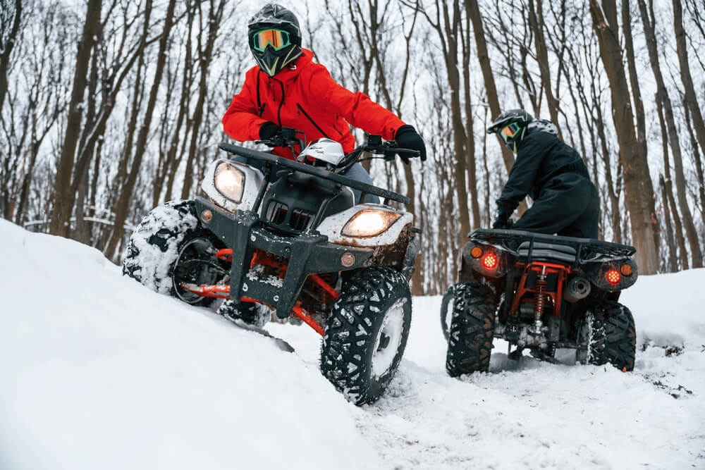 Two ATV riders wearing helmets and protective gear driving through snowy British Columbia trails, demonstrating essential ATV safety equipment.